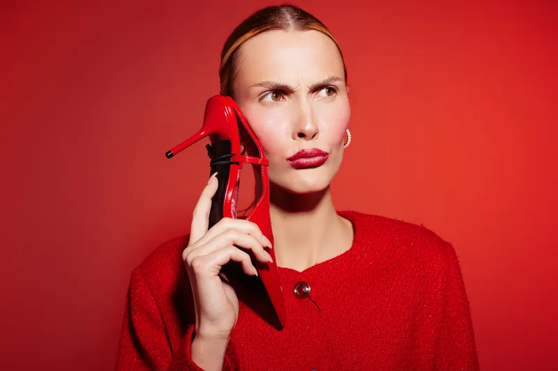 Woman in red sweater holds red high heel shoe near face with confused expression on red background. Concept for fashion, style, and unusual communication.