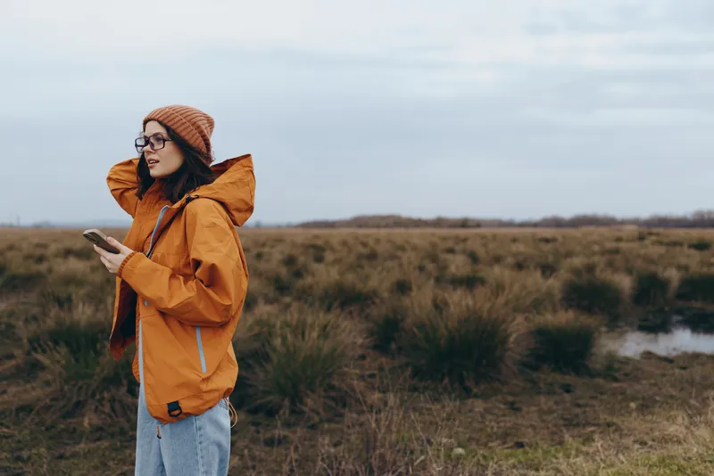 Woman outdoor lifestyle in orange jacket and beanie walking across landscape field Soft light nature scene with glasses and casual style Calm active travel mood