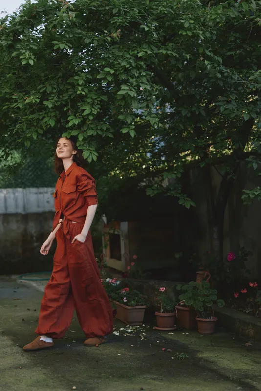 A woman in a rust red jumpsuit walks through a lush garden, sunlight on her face, embodying relaxed outdoor living and everyday style in a bright, candid moment