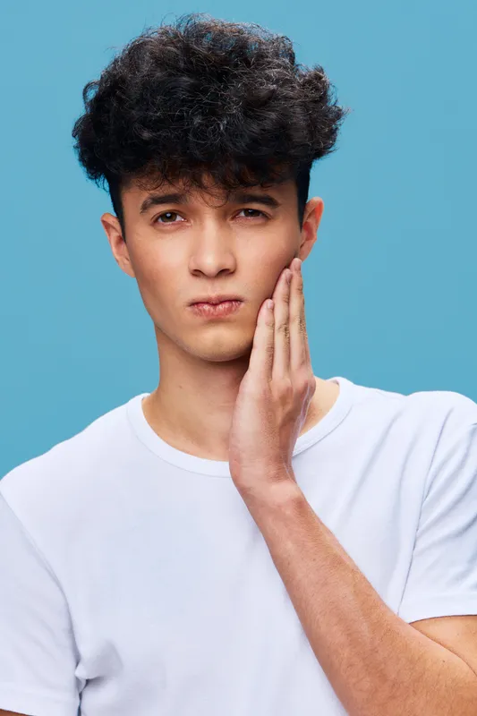 young man with toothache touching cheek in pain, wearing white t-shirt, curly hair, isolated on blue background, expressive facial expression showing discomfort and sensitivity