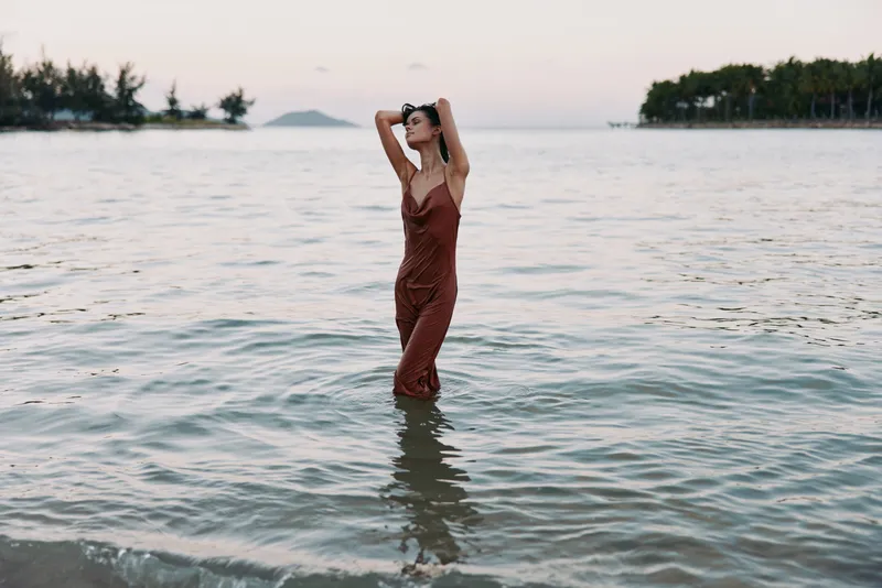 A woman stands waist deep in calm sea water at sunset, wearing a rust colored dress. She raises her arms, creating a graceful silhouette and soft reflective ripples.
