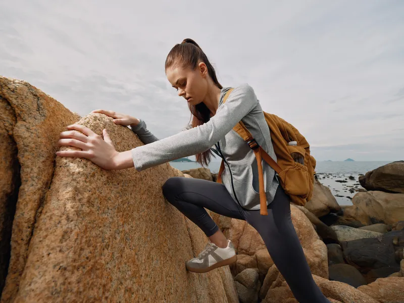 Young woman climbing rocks in outdoor adventure, wearing casual clothes and backpack, expressing focus and determination amidst a natural setting