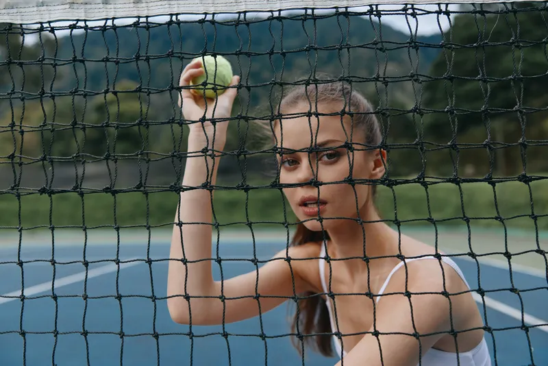 Athletic young woman holding a tennis ball, looking through a net on a tennis court, showcasing determination and focus in a vibrant summer setting