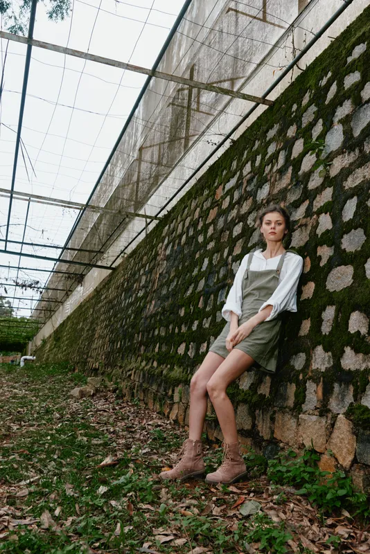 Stylish model in green overalls, showcasing confident expression against a rustic stone wall with nature elements for a trendy outdoor fashion vibe