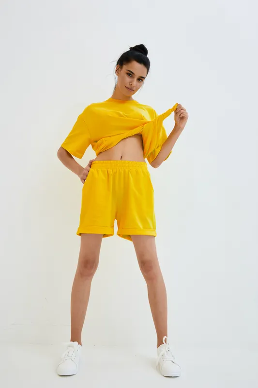 Young woman in bright yellow sportswear lifting her top, standing confidently against a white background, showcasing casual fitness style with white sneakers.