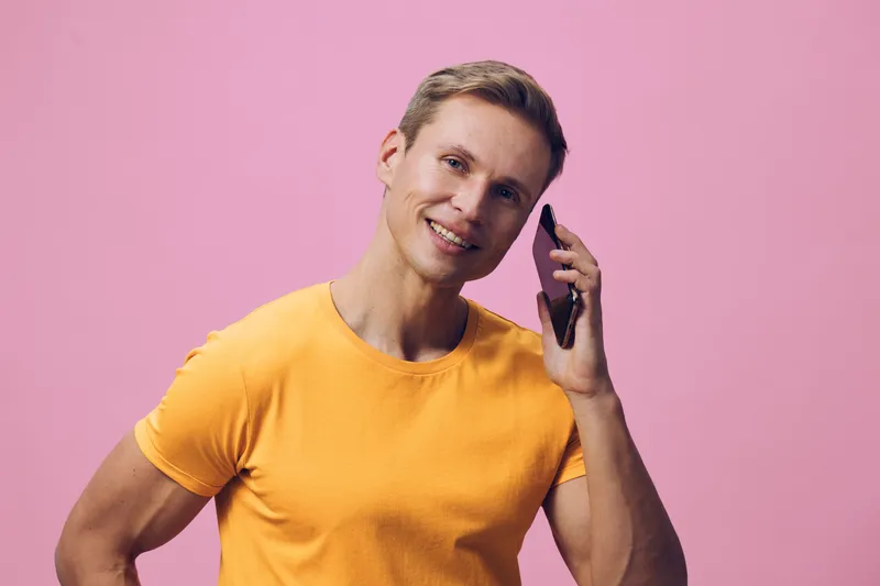 Smiling man wearing orange t-shirt talking on mobile phone isolated on pink background, casual lifestyle portrait, cheerful expression, relaxed pose, vibrant colors, modern style, positive mood.