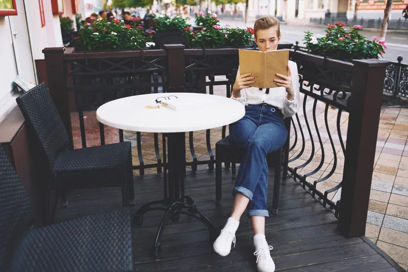 Стильная молодая женщина в рубашке сидит за столиком в летнем кафе с книгой в руках