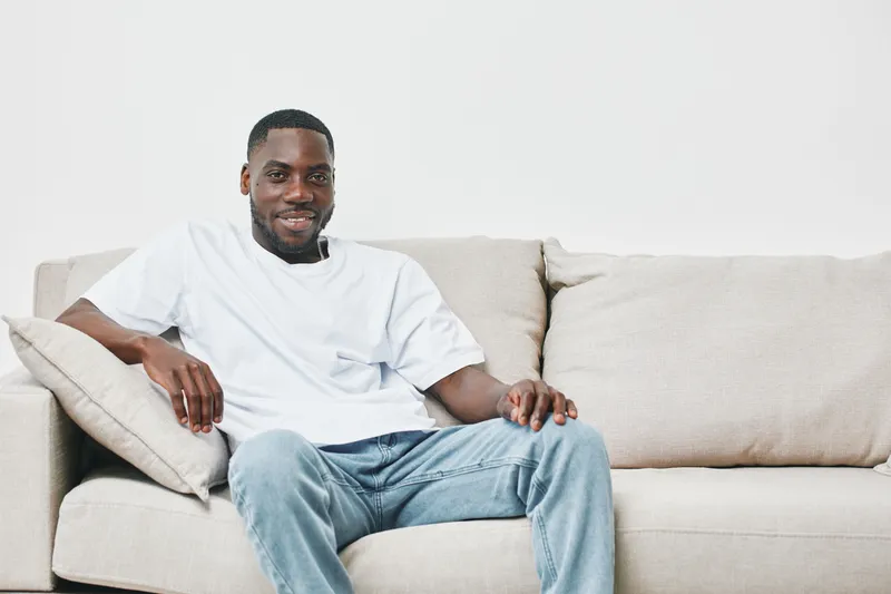 Smiling young Black man in casual white t shirt and jeans sitting comfortably on a light colored sofa, conveying relaxation and confidence in a modern setting