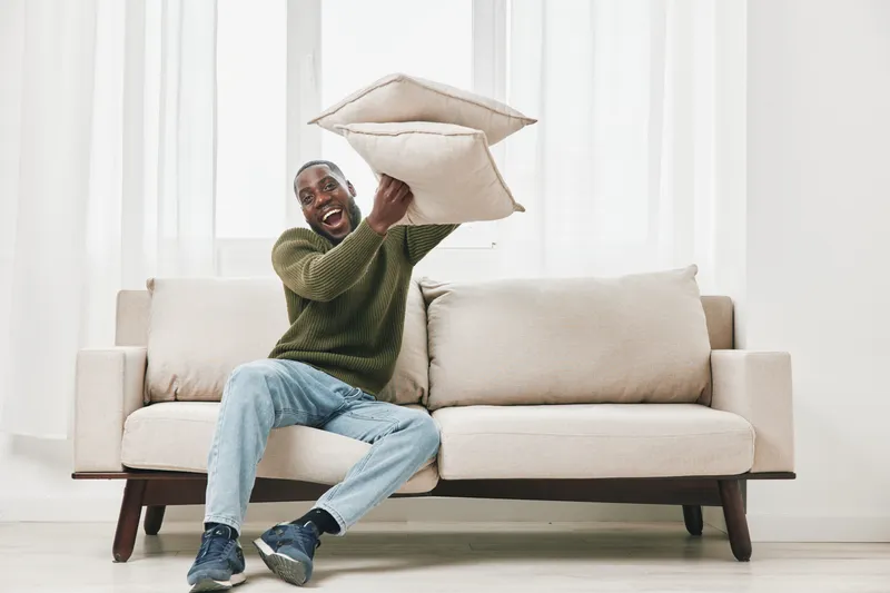 Happy man playfully throwing cushions on a light sofa in a bright room, showcasing joy and leisure, perfect for concepts of relaxation and comfort at home