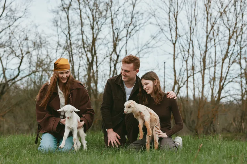 Smiling family outdoors with two goat kids, sitting on grass in a sunny field. Casual wardrobe, warm mood, playful moment, nature, countryside lifestyle, bonding and sunny mood. Cozy.