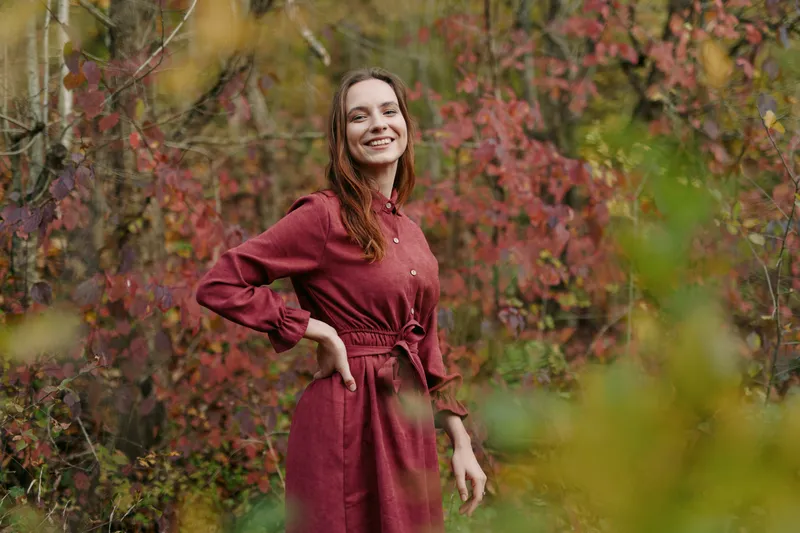 Smiling young woman in a simple red dress stands among autumn foliage, captured in an authentic natural moment that conveys genuine joy, warmth and subtle confidence in everyday realism.