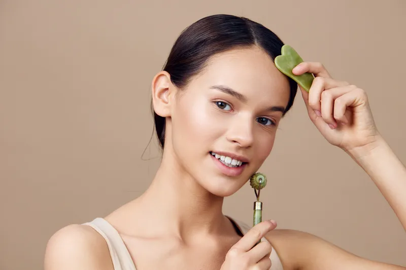 Young woman with smooth skin using jade roller and gua sha tool, promoting healthy skincare routine in a minimalist setting