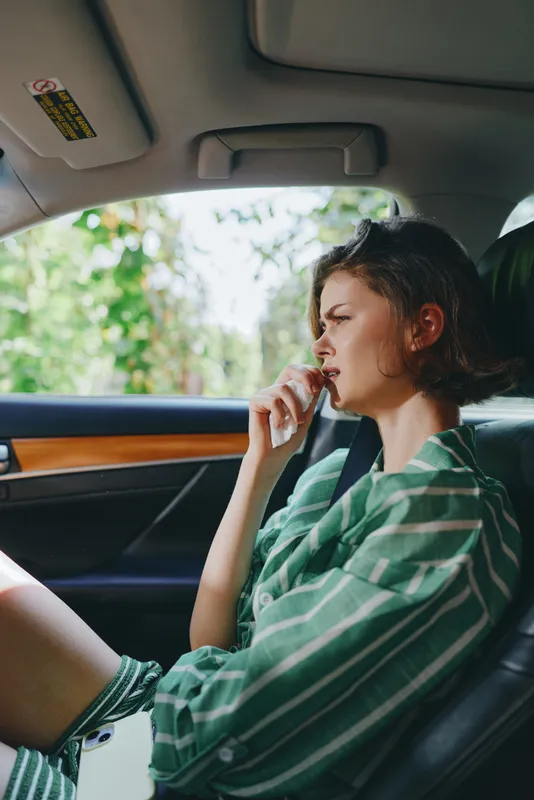 Young woman sits in a car interior, wearing a green striped shirt, looking thoughtful as soft light filters through the window.