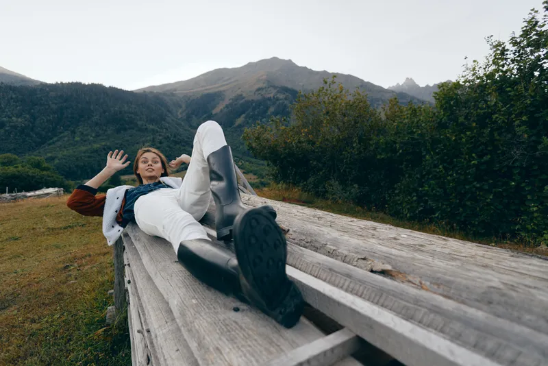 Woman in boots reclining on a wooden fence in mountains, nature meadow scene, autumn colors and relaxation moment. Casual coat and scarf, countryside leisure and scenic escape.