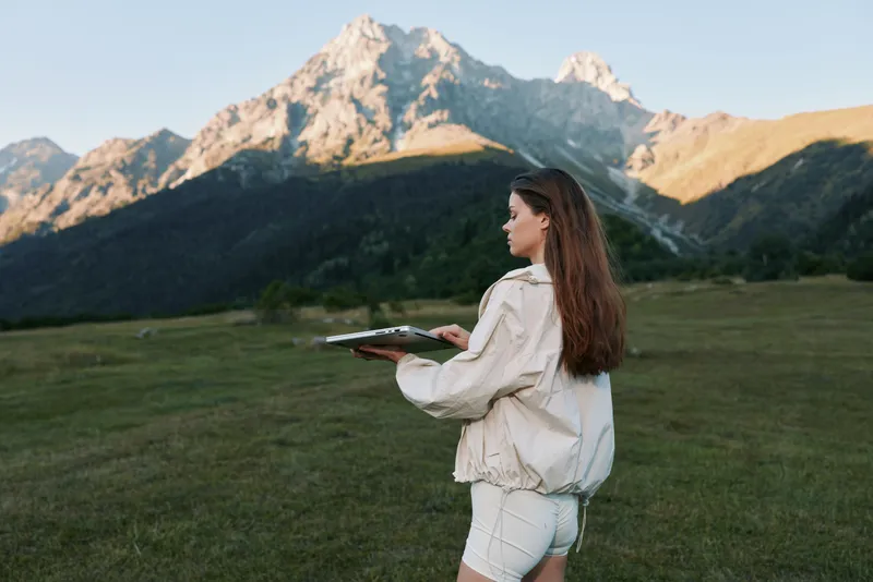 A young woman with long hair stands in a grassy field with a mountain range behind her, holding a clipboard and looking toward the horizon, capturing a calm outdoor scene.