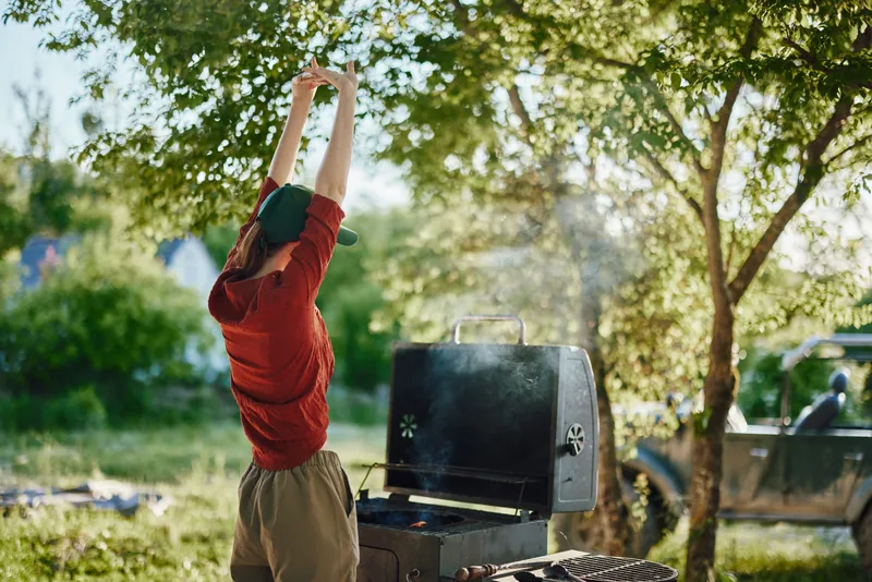 A young woman joyfully stretching in a casual red sweater and cap while grilling outdoors amid green nature, showcasing an active and vibrant lifestyle