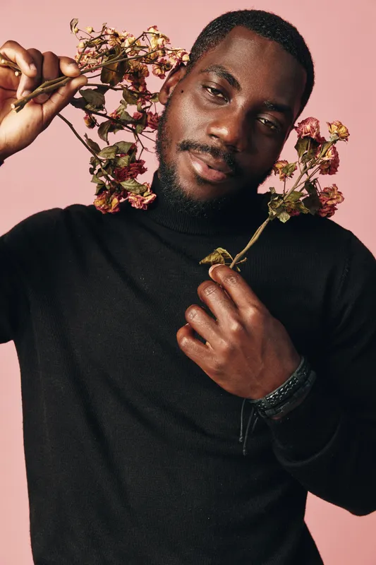 African man with dark skin wearing black sweater holding dried flowers near face in creative conceptual visual style with pink background and artistic expression.