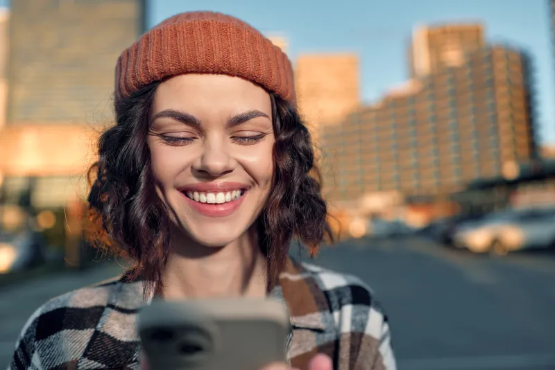 woman smile phone urban candid lifestyle authenticity golden glow: young woman smiling at smartphone in city street during golden hour, emotional storytelling and mindful living urban portrait.
