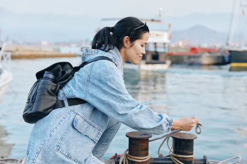 Harbor, woman, rope, dock, water, denim, backpack occupy frame as a lone figure inspects maritime equipment by a bollard, with boats and distant hills providing a calm harbor backdrop