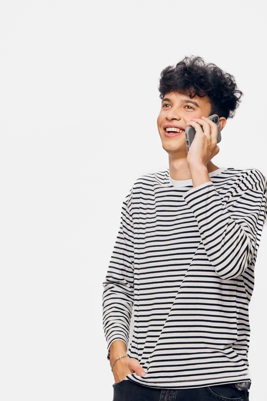young man talking on phone wearing striped long sleeve shirt smiling with joy isolated on white background, casual style and curly hair