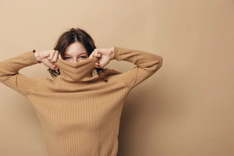 Young woman in brown knitted sweater pulls collar over face, peeking with eyes. Neutral beige background, casual style, cozy autumn or winter fashion concept with playful mood.