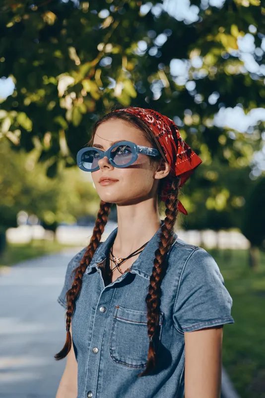 Lifestyle woman in modern boho-western denim street style with braided hair, blue sunglasses, and red bandana, captured in warm film color under sunny outdoor trees.