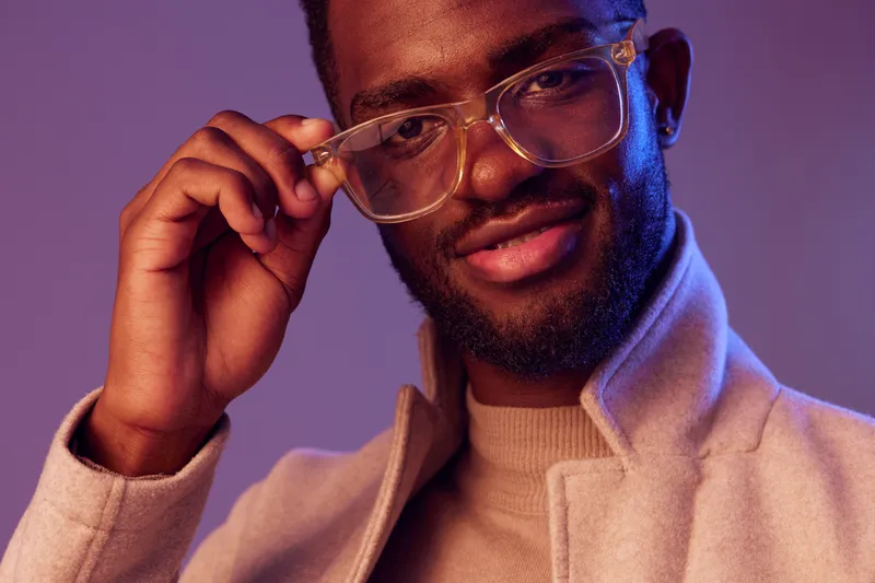 Stylish young African man in glasses smiling confidently against a vibrant purple background, demonstrating contemporary fashion trends
