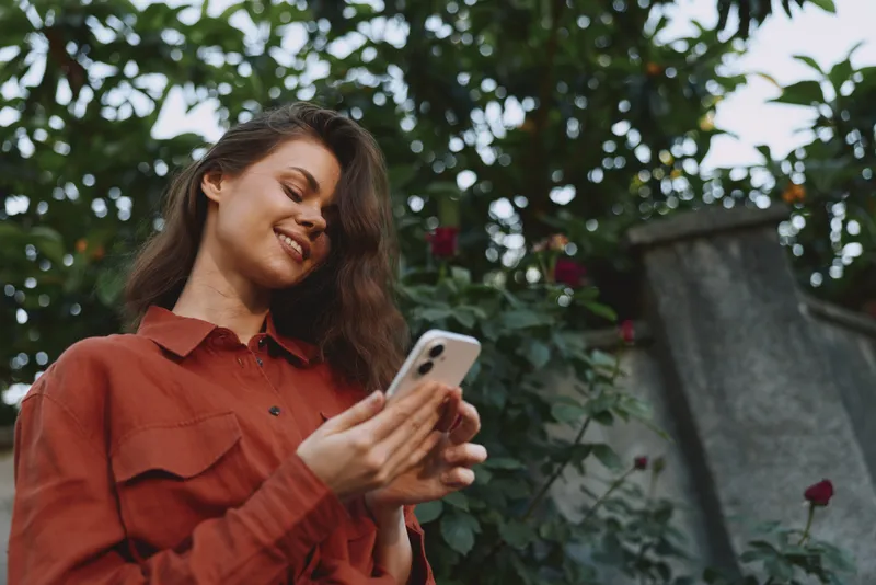 Young woman with brown hair smiles while checking her phone outdoors among lush greenery, conveying a casual, everyday moment