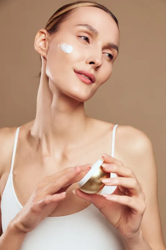 Young woman with pale skin and pulled-back hair applies white face cream on her cheek while holding a golden jar in a minimalistic studio setting with a beige background.
