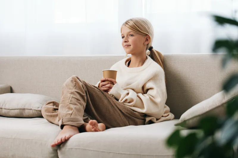 Authentic moment of a child relaxing at home after study, casual wardrobe, warm light, quiet lifestyle, cozy sofa, cup in hand, gentle game inspired mood.