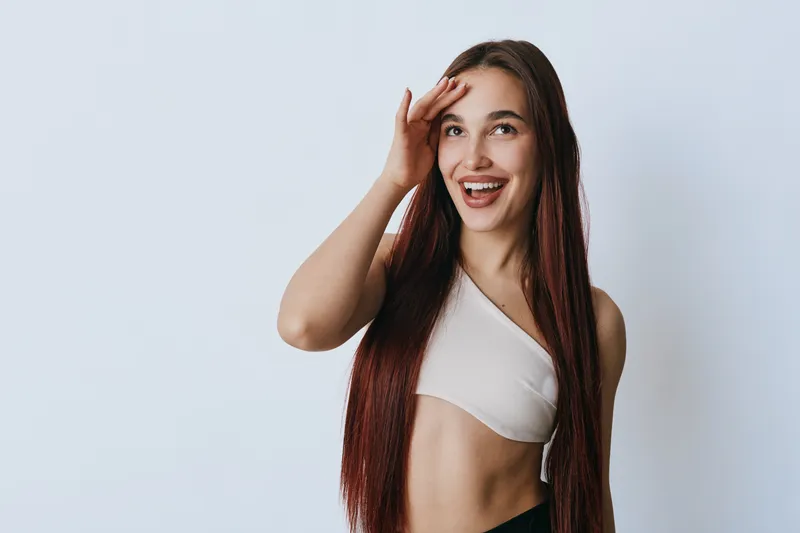 Young woman with long hair smiling and touching forehead posing in front of white background. Female model wearing sleeveless top showing happy expression and natural beauty.