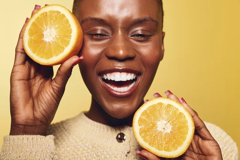 Happy African woman with dark skin holds two halves of fresh orange near face, smiling with closed eyes. Concept for healthy lifestyle, vitamin C, skincare, and natural beauty.