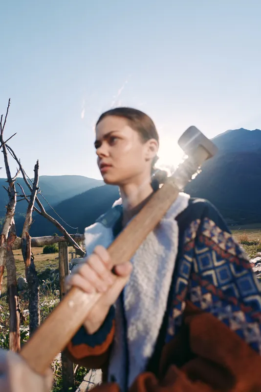 Woman hammer outdoors mountains portrait rural field sledgehammer, young laborer holding tool in backlit landscape, sunlight on patterned jacket, focused expression and workwear detail.