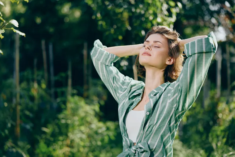 Woman outdoors, eyes closed, basking in sunlight with a striped green shirt, standing in a lush park, feeling calm and free.