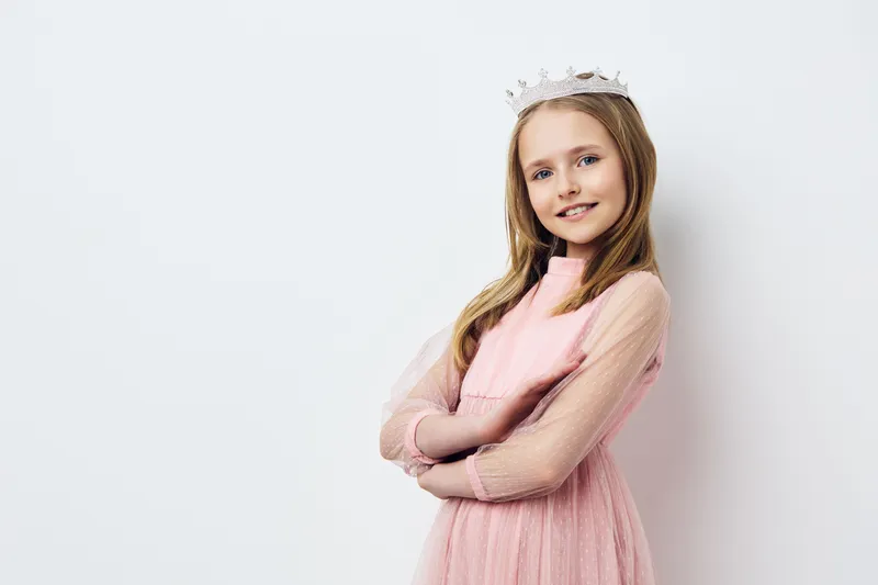 Young girl in a pink dress and crown, smiling confidently against a light background, exuding joy and creativity in a playful fashion