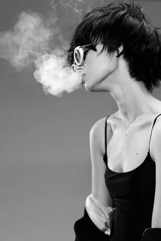 Black and white portrait of a stylish woman exhaling smoke with a calm expression in a dark outfit