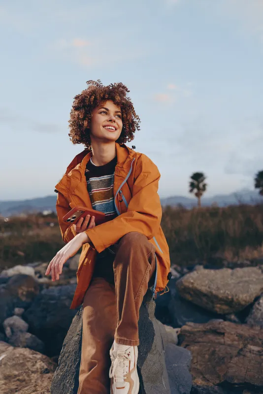 Woman lifestyle outdoors, smile, curly hair, orange jacket, sitting on rock in nature landscape, mountains and clouds behind, relaxed portrait, daylight adventure vibe, fresh air today
