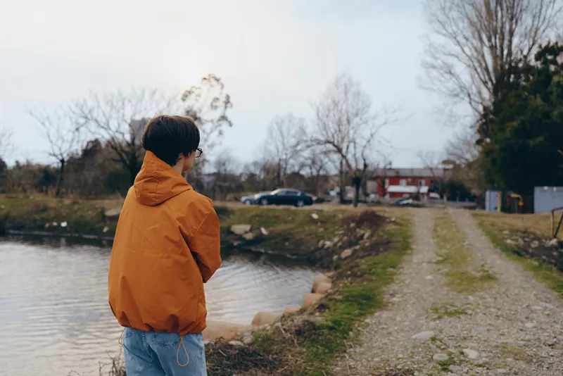 Woman in orange jacket and jeans stands by a lakeside path, outdoors lifestyle moment, looking at calm water, cloudy daylight, bare trees and distant walkway, quiet scenery, winter air
