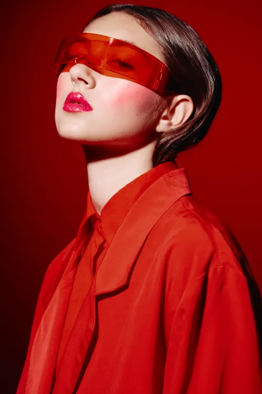 Fashion model in bold red outfit and tinted futuristic sunglasses poses with head tilted back. Studio shot with strong lighting on red background. Concept of beauty fashion creativity.