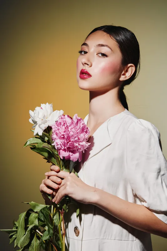 Young woman in stylish white outfit posing with pink and white peonies. Beauty fashion creative portrait on yellow background emphasizes elegance and floral charm.