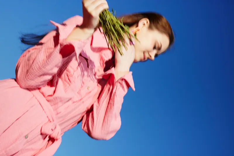 Young woman in a pink coat holding fresh herbs against a clear blue sky, exuding joy and confidence, representing healthy eating and a vibrant lifestyle