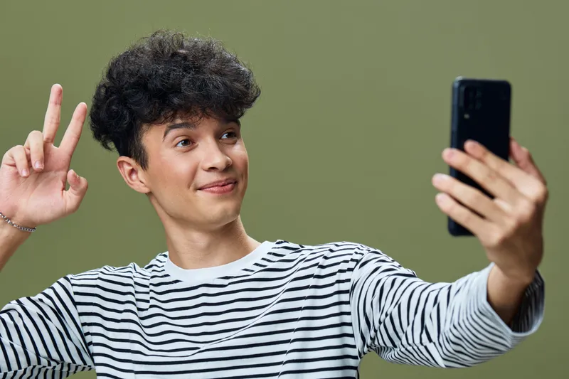 Young man taking a selfie with peace sign, wearing a striped shirt, smiling happily against a plain olive green background, casual and cheerful mood.