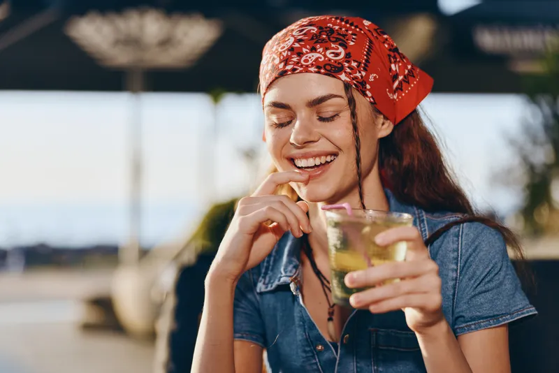 Lifestyle woman wearing modern boho-western denim street style with a red bandana, smiling while holding a refreshing drink in film color tones.