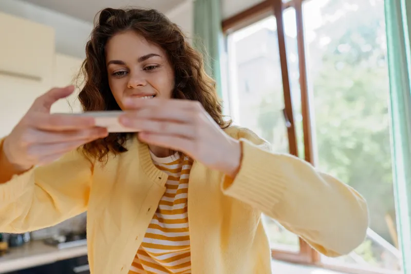 Young woman with curly brown hair wearing a yellow cardigan takes a casual selfie at home, smiling softly as she captures a moment of everyday life.