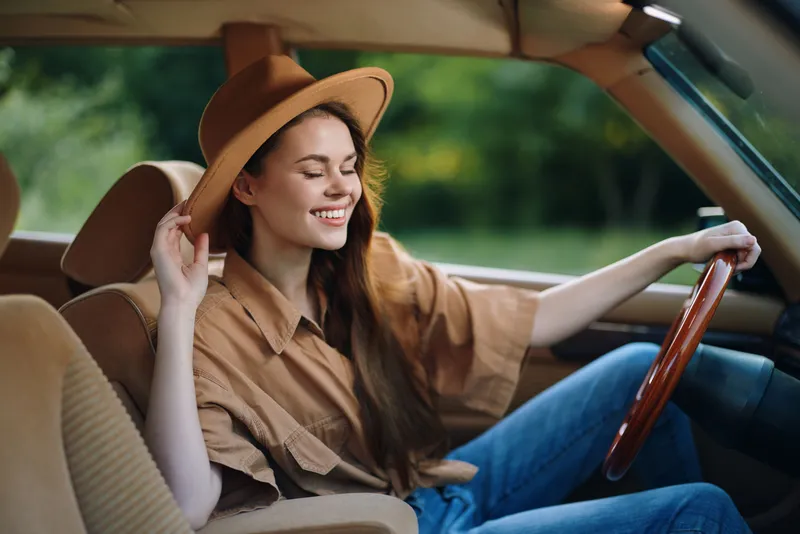 Young woman enjoying a sunny day in a vintage car, wearing a wide-brimmed hat and casual clothing, exuding joy and confidence in a lush green background.
