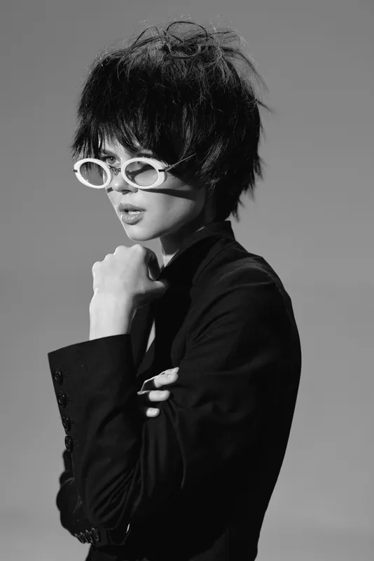 Black and white portrait of a stylish woman with short hair and white sunglasses wearing a dark blazer in a minimalistic setting