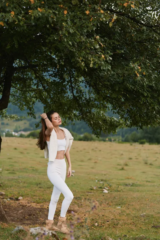 Young woman dancing in a white outfit in an open meadow beneath a green canopy, captured in warm sunlit light, with her carefree pose conveying a relaxed joyful mood.