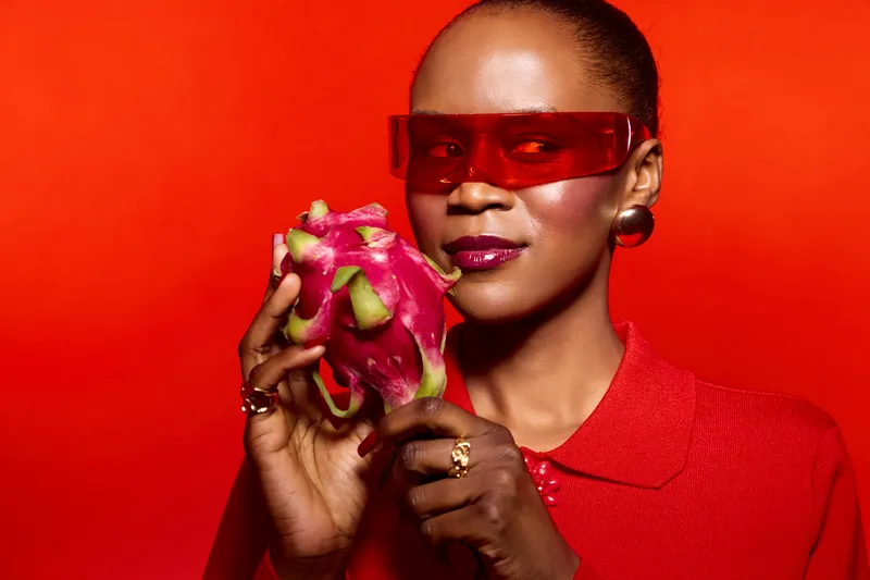 Stylish African woman with dark skin wearing red glasses and red shirt holds fresh dragon fruit against a vibrant red background. Concept of fashion, fruit, and bold colors.