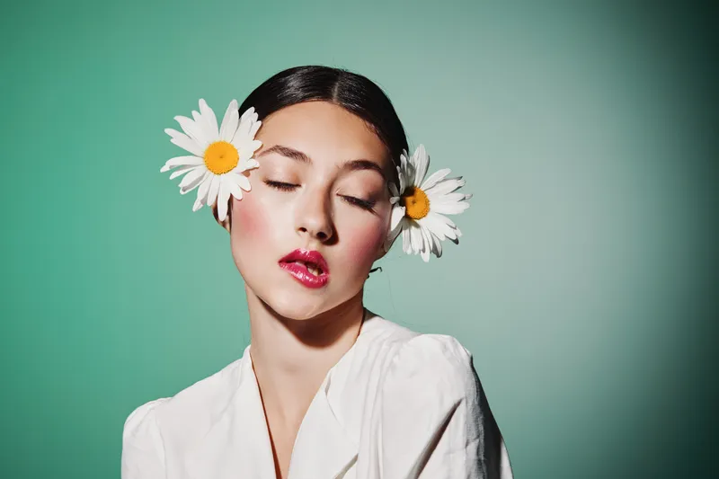 Fashion young woman with natural makeup and flowers in hair posed with closed eyes on green background. Concept for beauty, creative, and fashion editorial.