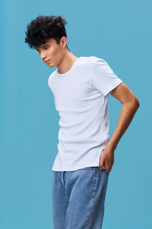 Young man with curly hair wearing plain white t-shirt and blue jeans stands against blue background, looking down thoughtfully in casual style portrait.